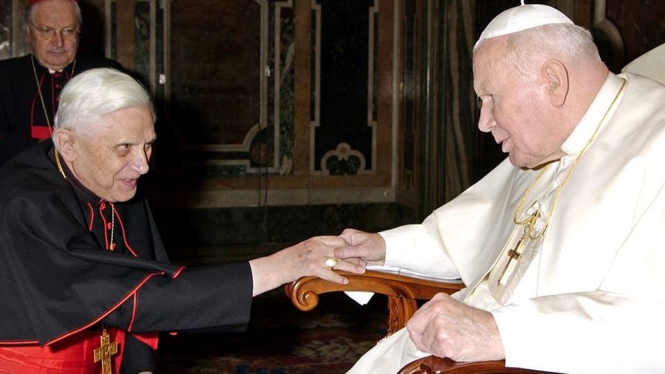 Papst in weissem Umhang auf einem Stuhl, Ratzinger in Kardinalskleidung reicht ihm die Hand