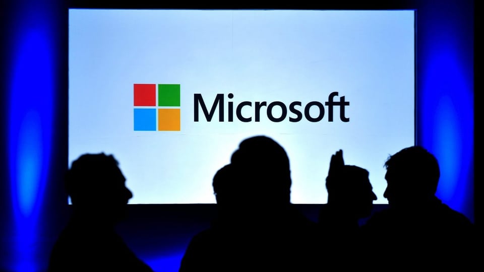 Microsoft-Logo auf einem Bildschirm, einige Menschen davor.