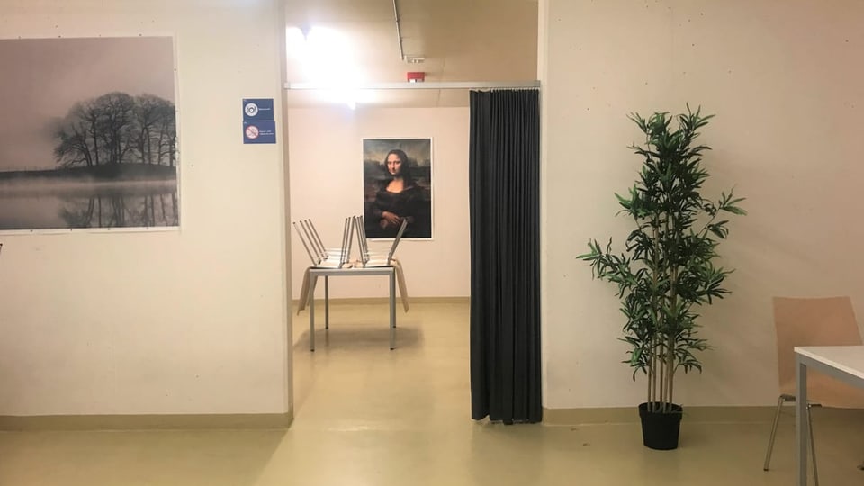 Kahler Raum mit Bildern an den Wänden, eine künstliche Pflanze, ein paar Tische und Stühle