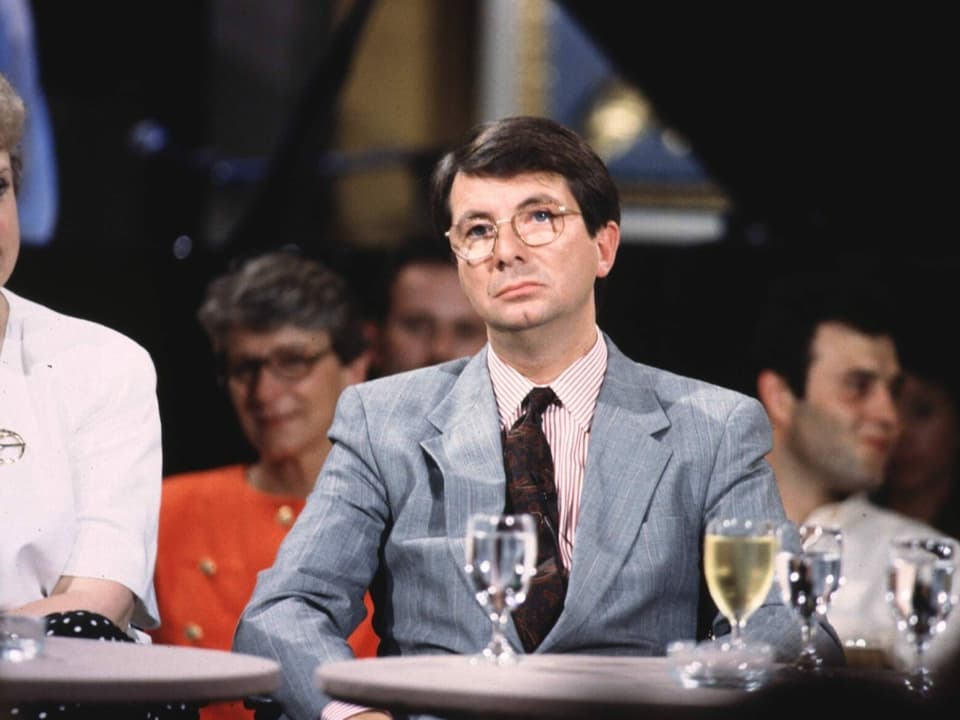 Mann mit Brille und Anzug im Stil der 1990er-Jahren sitzt an einem Tisch. Vor ihm ein Wasserglas.