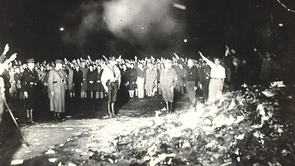 Schwarz-weiss Foto von einem Haufen lodernder Bücher vorne, im Hintergrund Schaulustige, die den Hitlergruss machen.