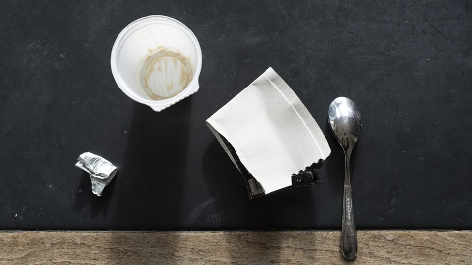 Ein leergegessener Joghurtbecher steht auf einem Tisch. Daneben die Alufolie, der Kartonmantel und ein Löffel.