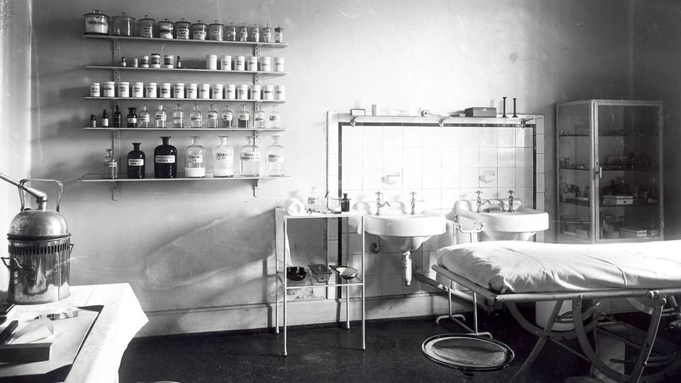 Historisches Bild: Behandlungszimmer mit Liege und Gestellt mit Medikamenten.