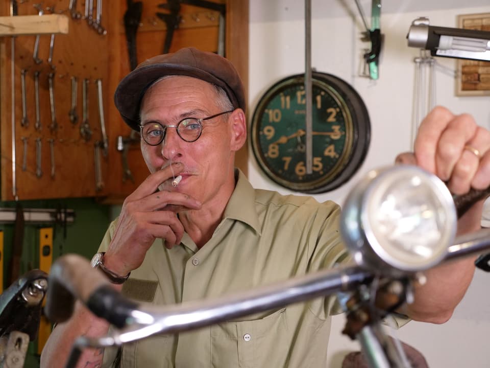 Ein rauchender Mann mit Mütze, seine linke Hand lehnt lässig auf einem alten Fahrradlenker. 