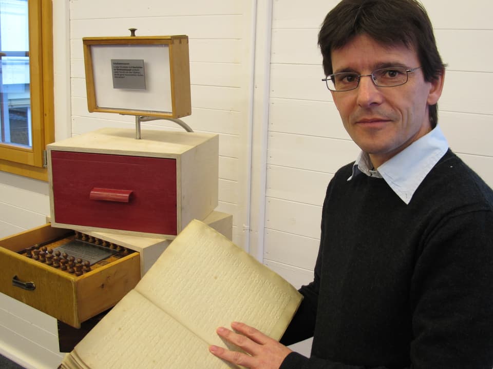 Christian Niederhauser neben Schubladen: in einer liegt ein Schachspiel, ein altes Buch hält er offen in der Hand.