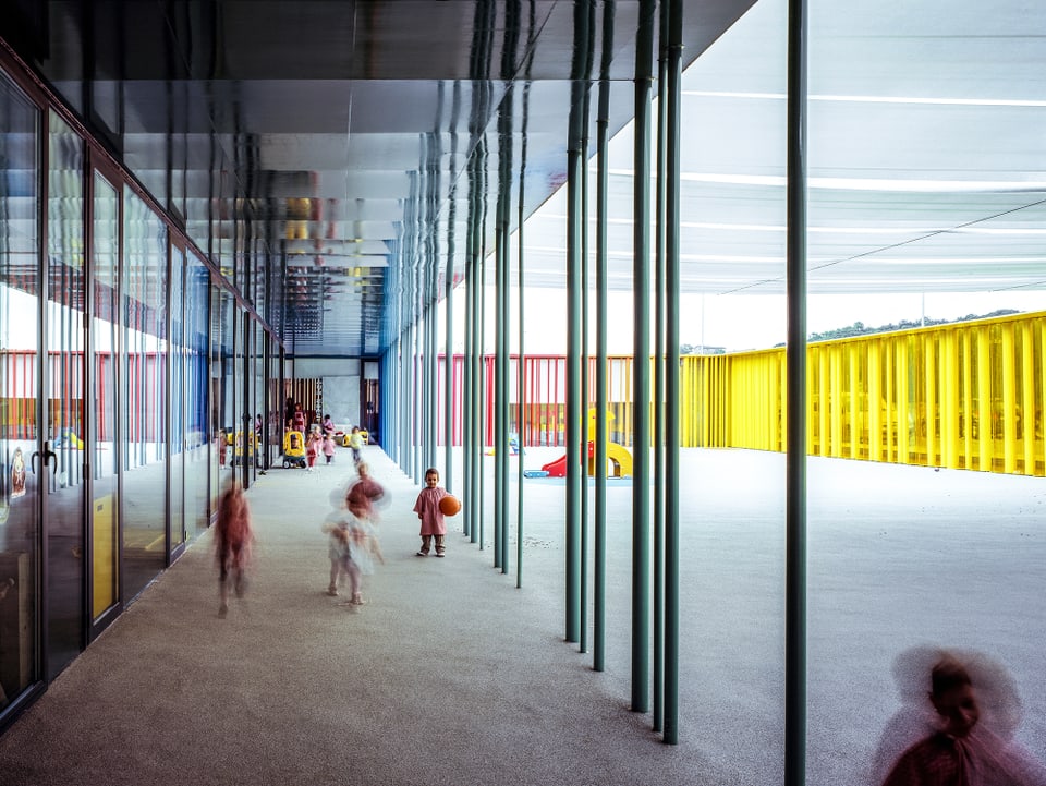 Ein gelb und rot rahmter Innenraum eines Kindergartens, der von grünen Säulen halbiert wird. Darin spielen Kinder.