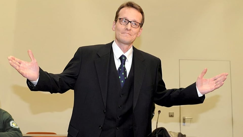 Mann mit braunen, kurzen Haaren gestikuliert mit einem Lächeln im schwarzen Anzug, trägt Brille.