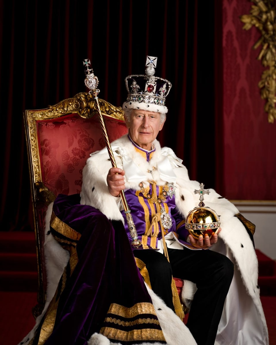 König Charles III. im vollen Ornat auf einem Thron im Buckingham-Palast sitzend.