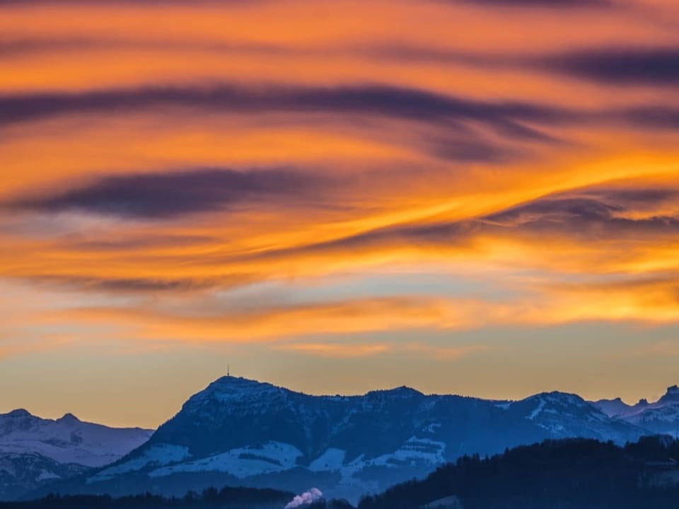 Wellige Wolken am Himmel, die rot leuchten. Unten Alpenpanorama mit etwas Schnee. 