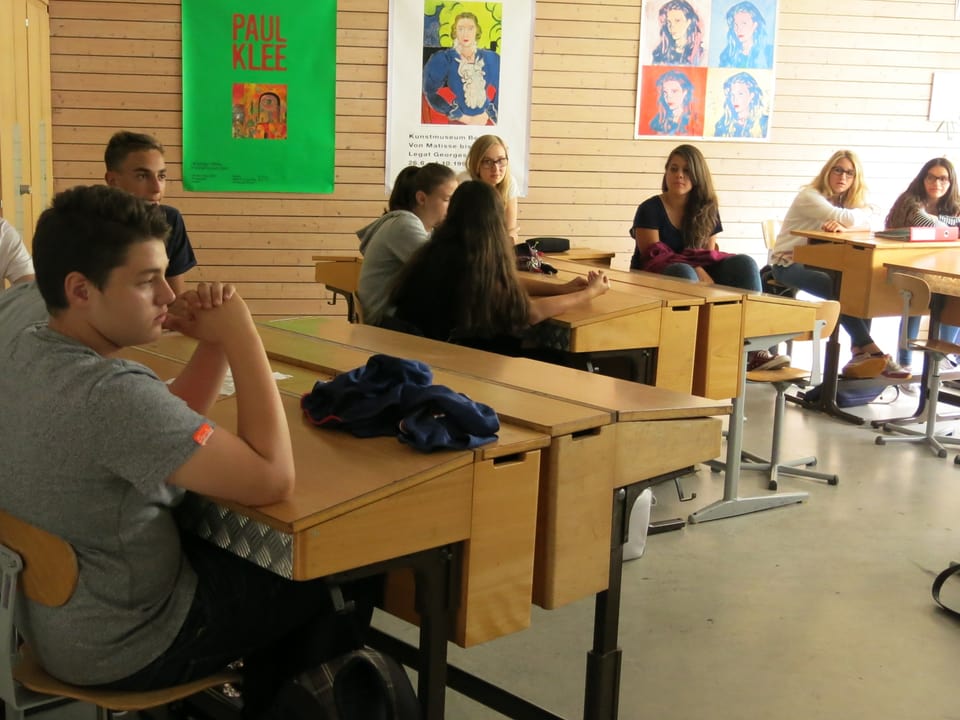 Schüler sitzen an Pulten und schauen in verschiedene Richtungen. 