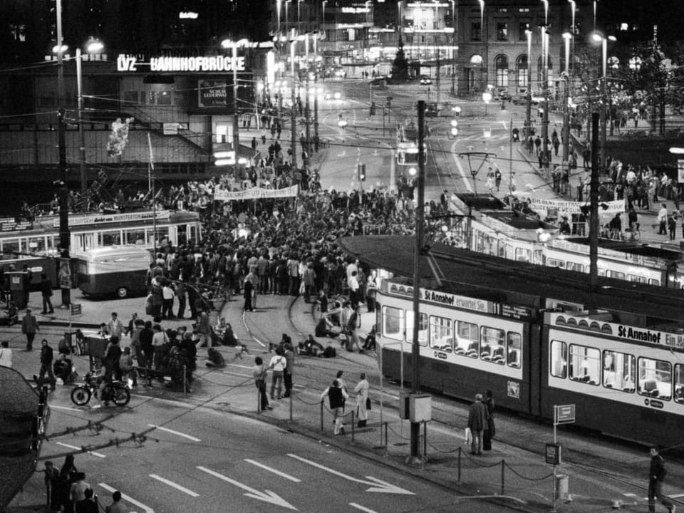 Belebter Strassenbahnhof bei Nacht mit Menschen und Strassenbahnen.