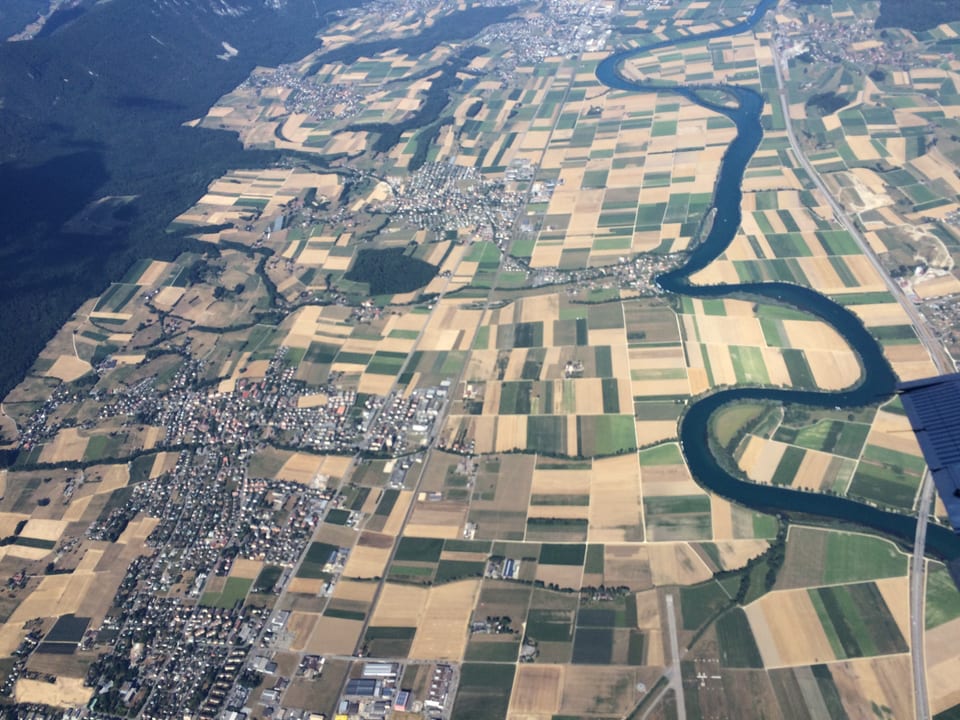Flugbild aus grosser Höhe auf das Schweizer Mittelland. Mit der Trockenheit sind die Felder hellbraun und sehr hell. In der Mitte schlängelt sich die Aare als blauer Schlauch durch die Felder. Die Felder sind schachbrettartig angeordnet. 