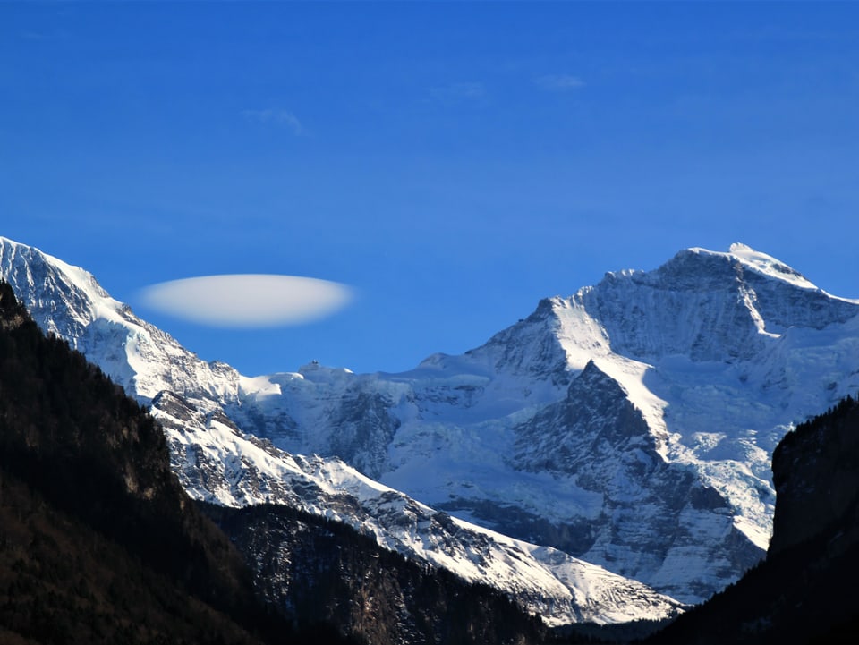Die Berner Riesen trohnen in weisser Pracht. Über dem Sattel des Jungfraujochs ist eine linsenförmige Wolke zu sehen.