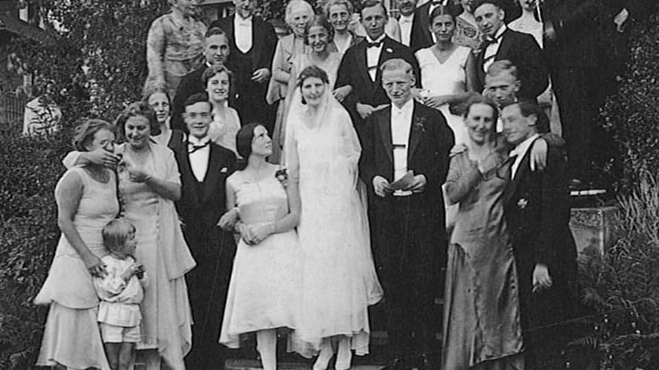 schwarzweiss Foto Hochzeitsgäste, Frau in weissem Kleid und Schleier, rechts Mann im Wrack und gegelten Haaren