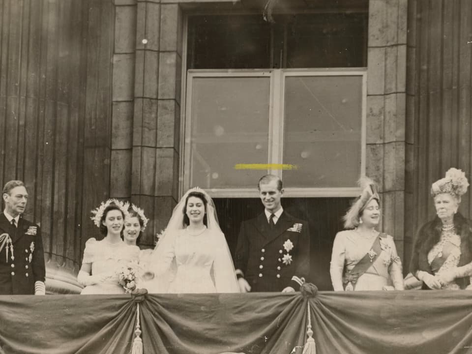 Menschen stehen auf einem Balkon. In der Mitte das Ehepaar, sie im Hochzeitskleid, er im Anzug.
