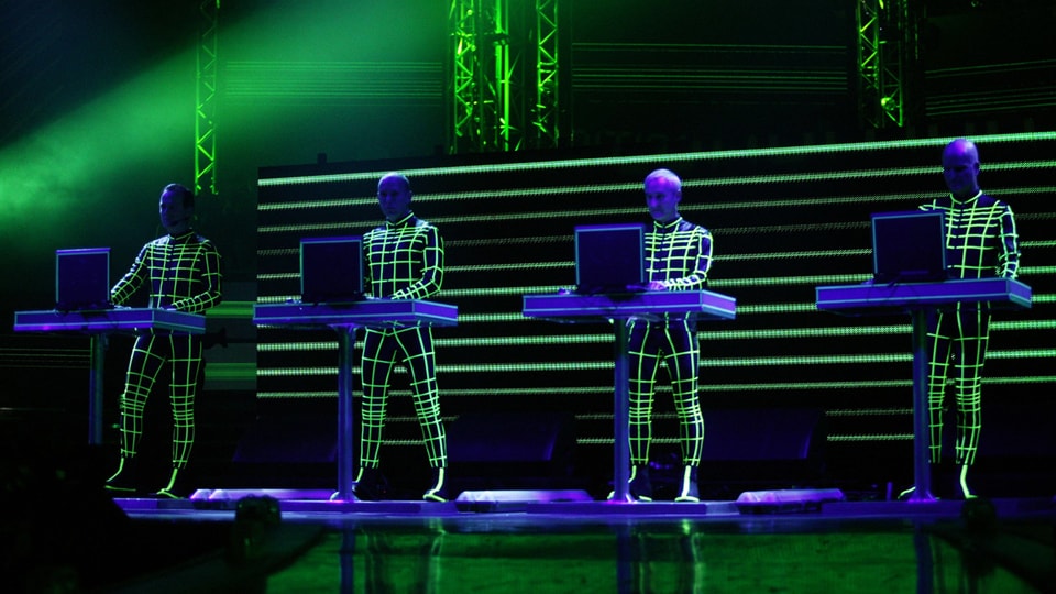 Vier Männer auf der Bühne, in schwarzen Overalls, die mit grün-blauem Licht bestrahlt werden.