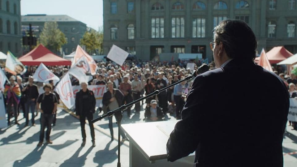 Rechts mit dem Rücken zur Kamera ein Redner im Anzug, steht am Rednerpult. Dahinter verschwommen protestierende Masse.