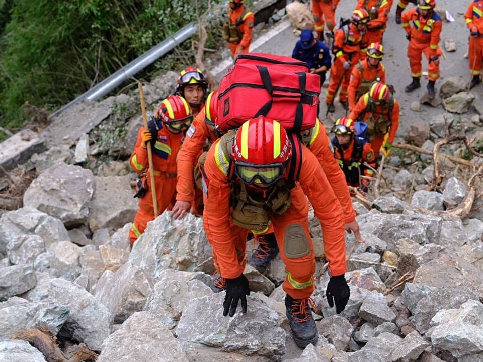Rettungskräfte steigen über einen grossen Berg von Steinen. Sie tragen rote Rucksäcke, Helme und orangene Overalls. Es sind rund 15 Personen auf dem Bild.