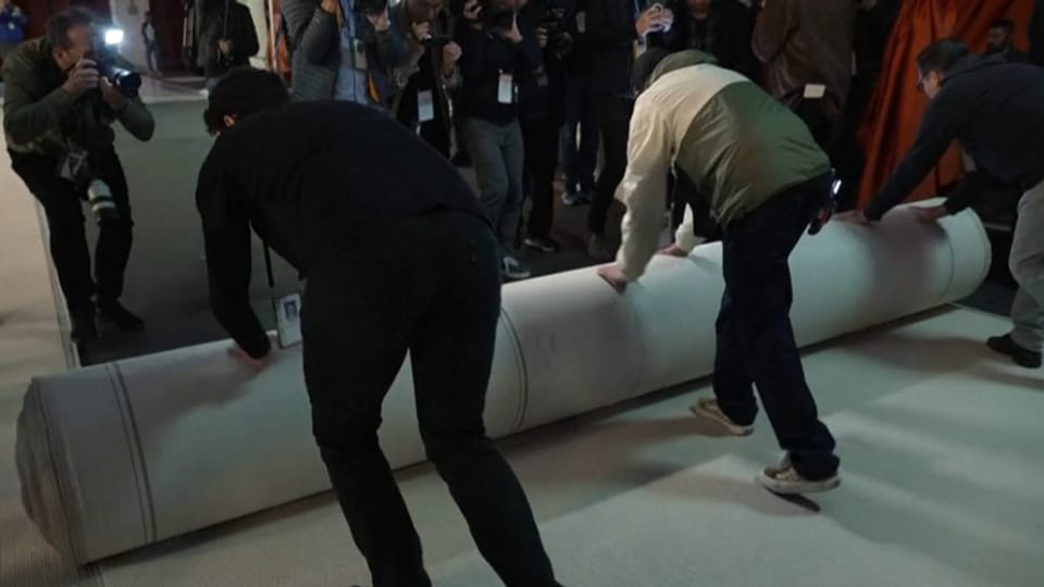 Zwei Männer rollen den beigen Oscar-Teppich aus. Rundherum fangen Fotografen den Moment auf Kamera ein.