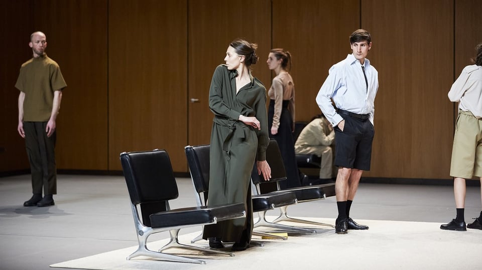A dozen men and women stand awkwardly around some chairs. Everyone looks in different directions.