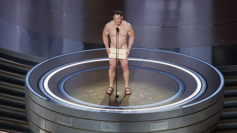 Der Wrestler John Cena steht nackt auf der Bühne, bedeckt nur mit einem Schild mit dem Wort «Costume design» drauf.