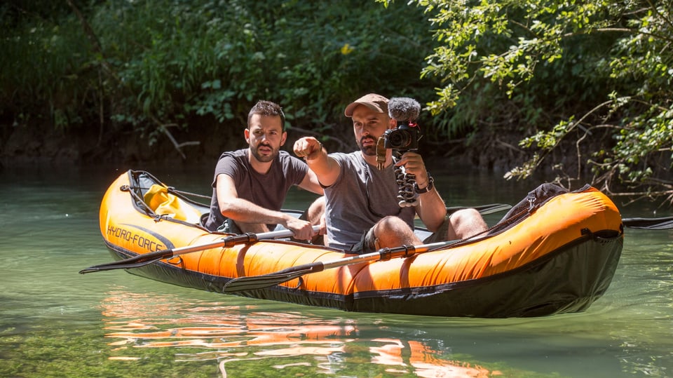 Julien Donzé hinten und Fabien Wohlschlag vorne sitzen in einem orangen Schlauchboot auf einem Fluss. Fabien Wolschlag hält eine Kamera in der Hand. 