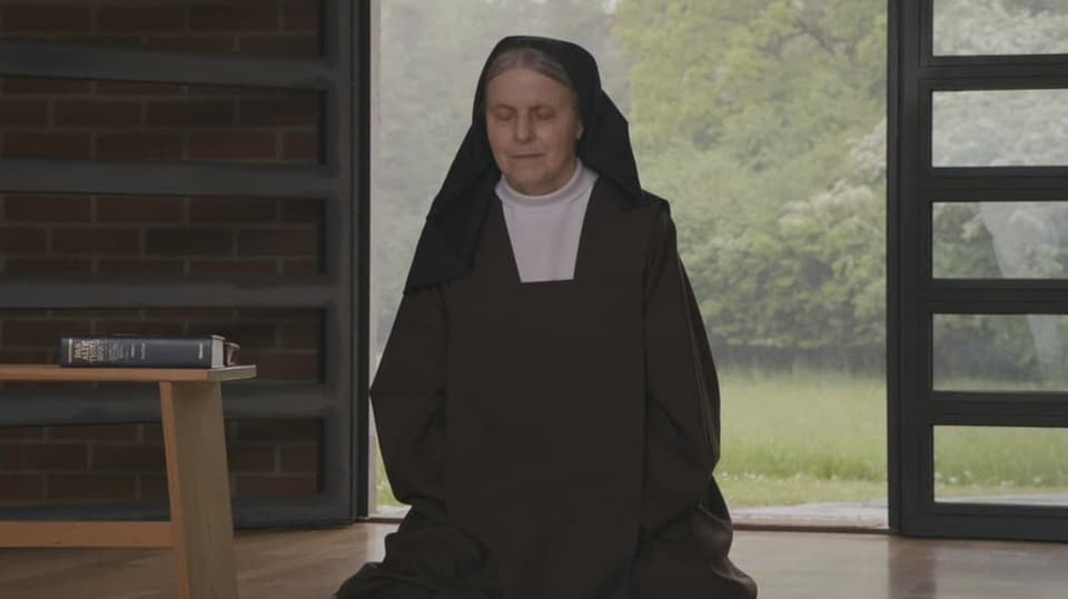 Eine Frau in schwarzem Gewand sitzt auf einem weissen Kissen und betet mit geschlossenen Augen.