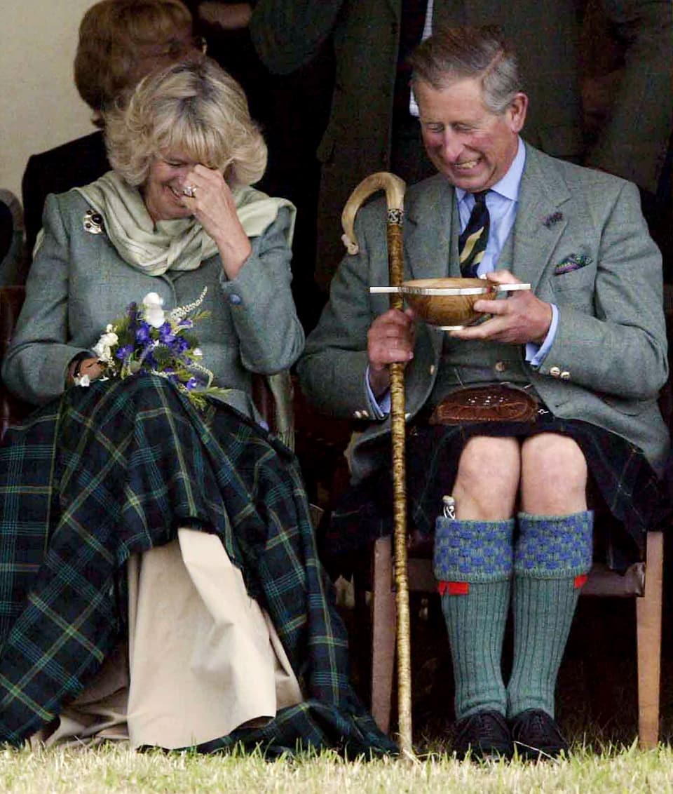 Charles und Camilla sitzen nebeneinander und lachen. Charles hält eine Holzschale in der Hand.