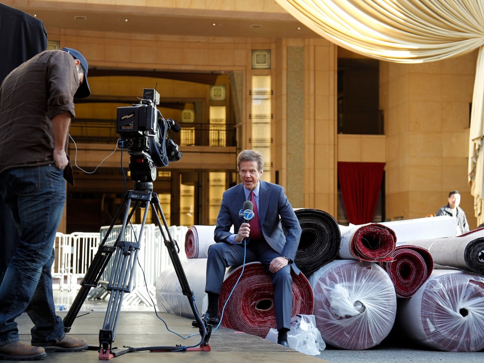 Ein Reporter sitzt auf zusammengerollten Teppichen. Er wird von einem Kameramann gefilmt.