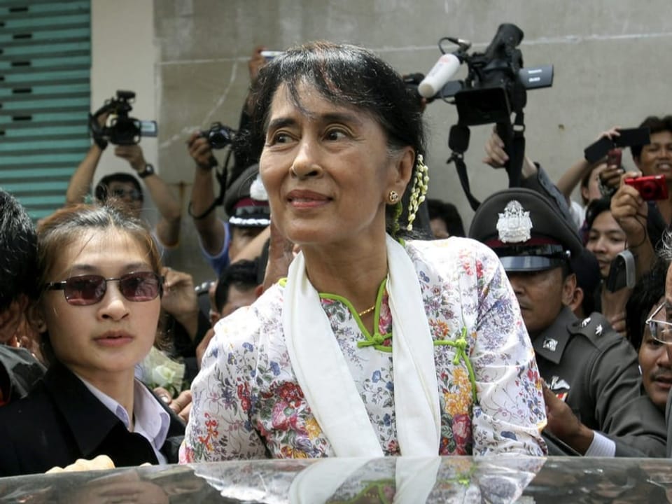 Aung San Suu Kyi steht vor einem Auto. Um sie herum stehen viele Leute und Fotografen. 