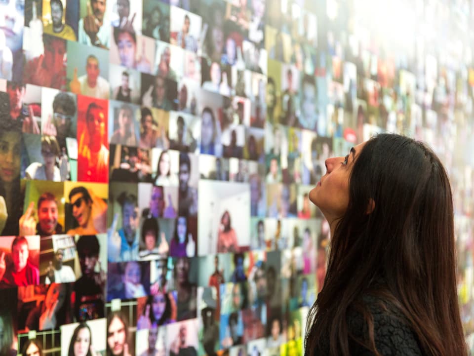 Eine Frau steht vor einer Collage, die aus Portraits von Menschen besteht, die ihren Mittelfinger in die Kamera halten.