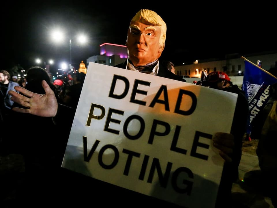 Eine Trumpmaske hält ein Schild mit der Aufschrift Dead People Voting in der Hand.