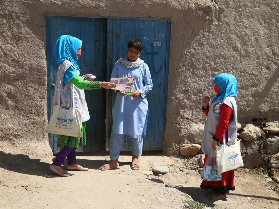 Zwei Frauen bringen einem Mann vor seiner Haustüre Informationsbroschüren. Die Frauen tragen ein hellblaues Kopftuch, was die Farbe von UNICEF symbolisiert.