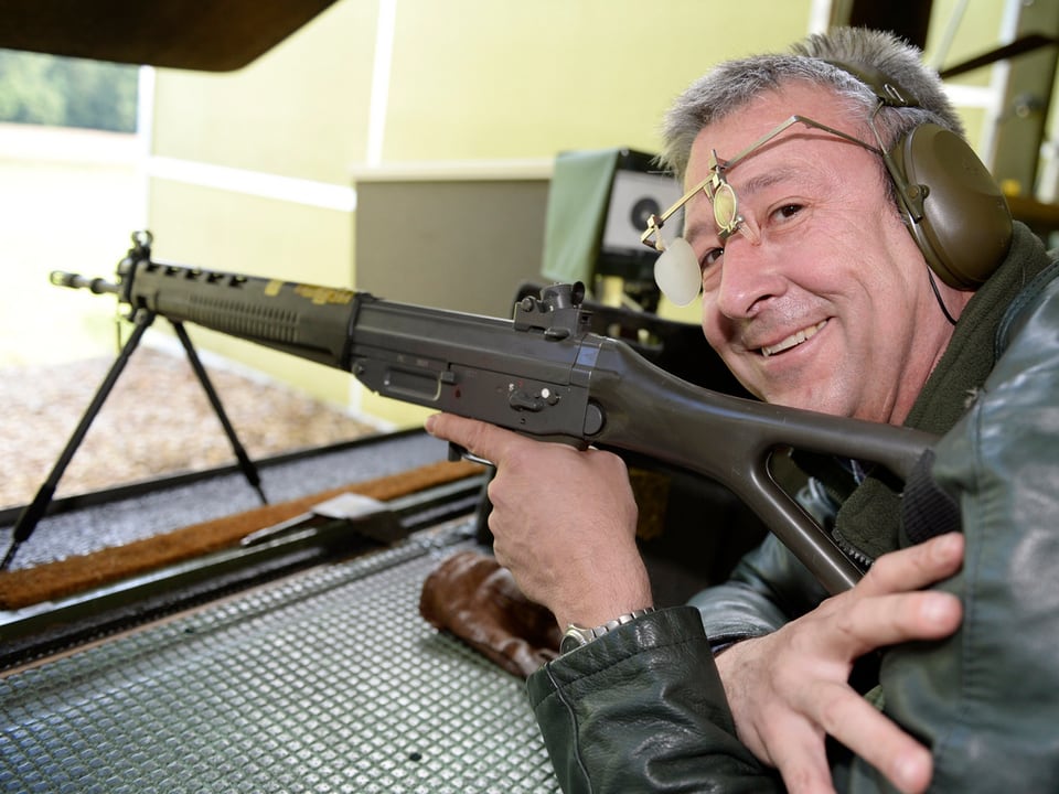 Ein Mann liegt auf dem Bauch, ein Sturmgewehr im Ansatz, und lächelt in die Kamera.