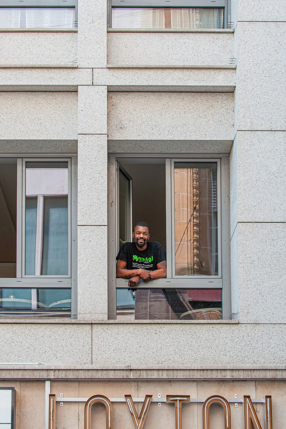 Ein junger Mann lehnt aus einem Fenster, darunter ein Leuchtschriftzug.