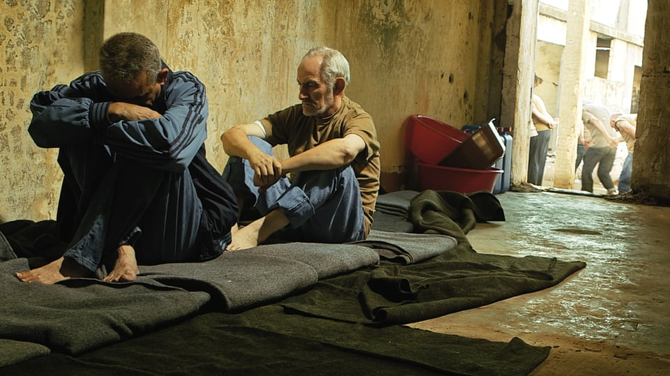 Zwei Männer sitzen mit gesenktem Kopf in einem Raum auf Wolldecken.
