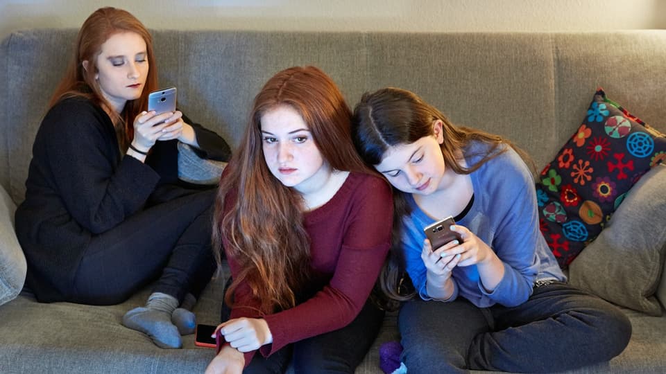 Drei Mädchen sitzen auf einem Sofa. Zwei von ihnen schauen auf ihr Handy. 