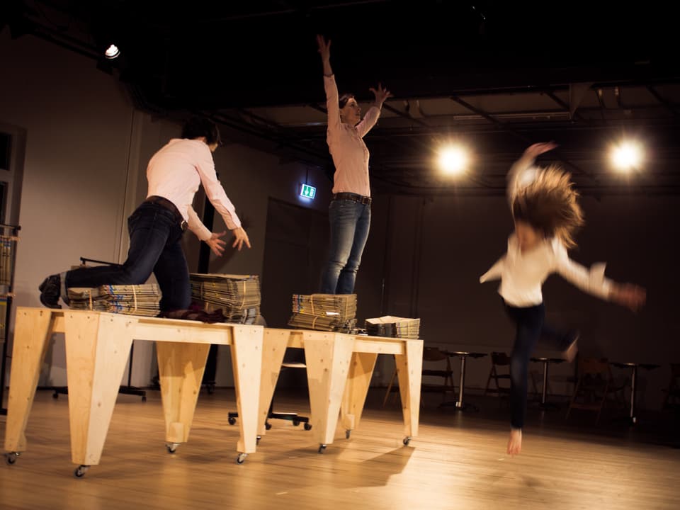 Drei Frauen auf einer Theaterbühne. Zwei davon knien und stehen auf Holztischen.