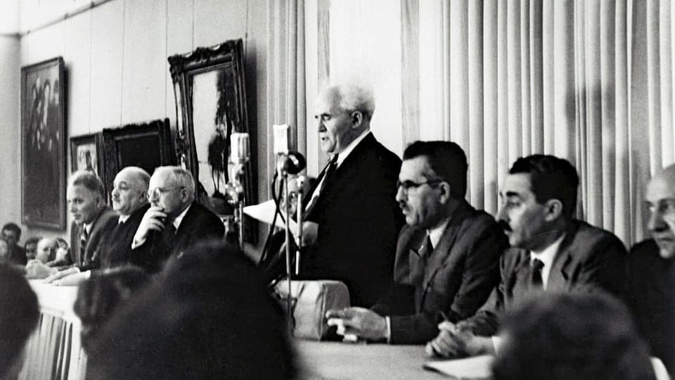 Gurion liest vor, am Tisch sitzen Männer.