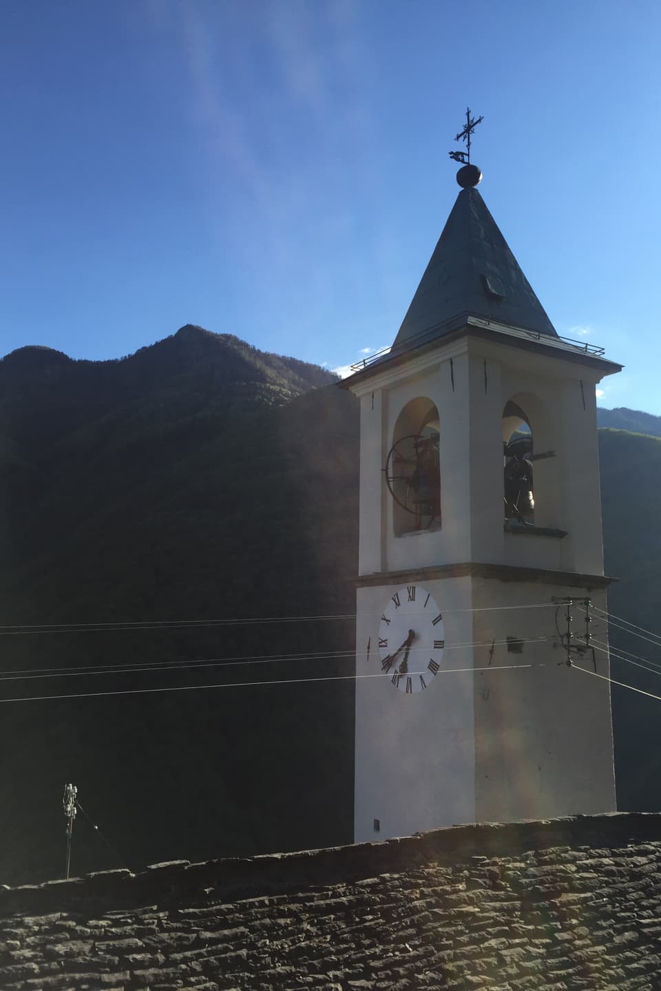 Alter Tessiner Kirchturm, die Uhr zeigt 20 vor sieben. Morgensonne. Im Hintergrund bewaldete Berge, im Vordergrund ein Steindach. 