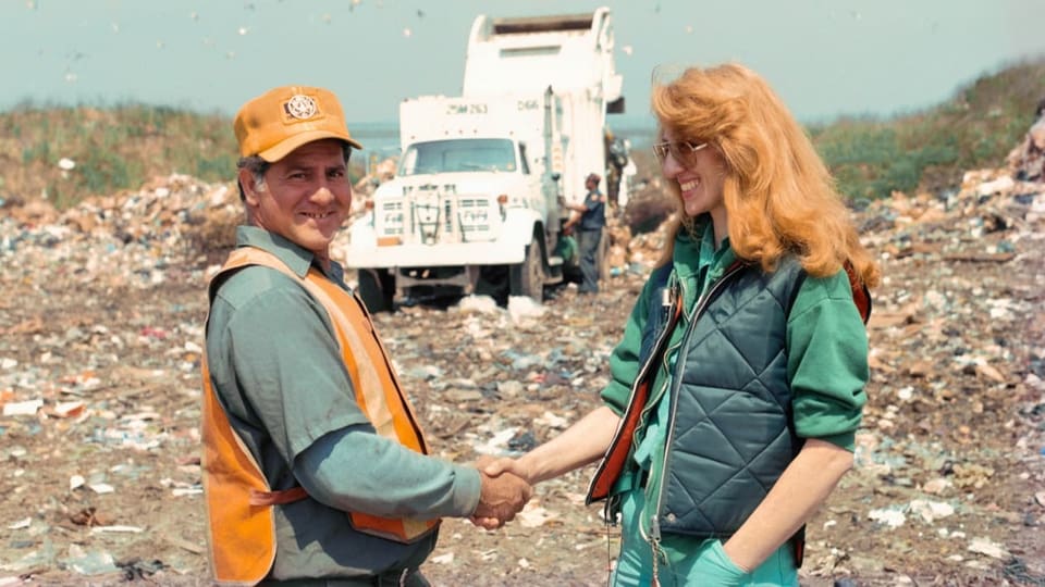 Mittdreissigerin mit roten langen Haaren schüttelt einem Müllmann die Hand. Sie stehen auf einer Müllhalde und lächeln.