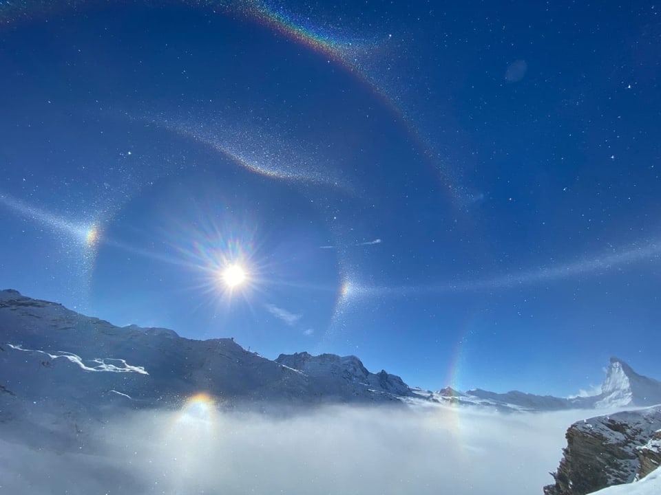 Verschiedene Haloeffekte mit der Sonne im Zentrum vor blauem Himmel. Dazu verschneite Berglandschaft.