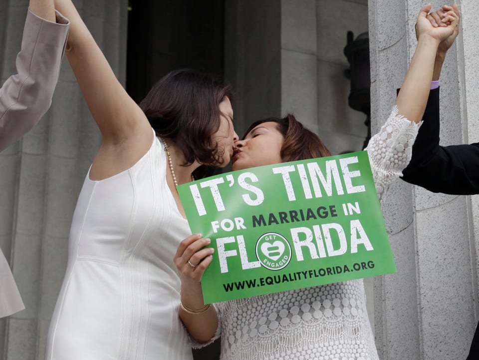 Ein lesbischen Paar steht vor einem Gebäude und küsst sich. Beide tragen weisse Hochzeitskleider.