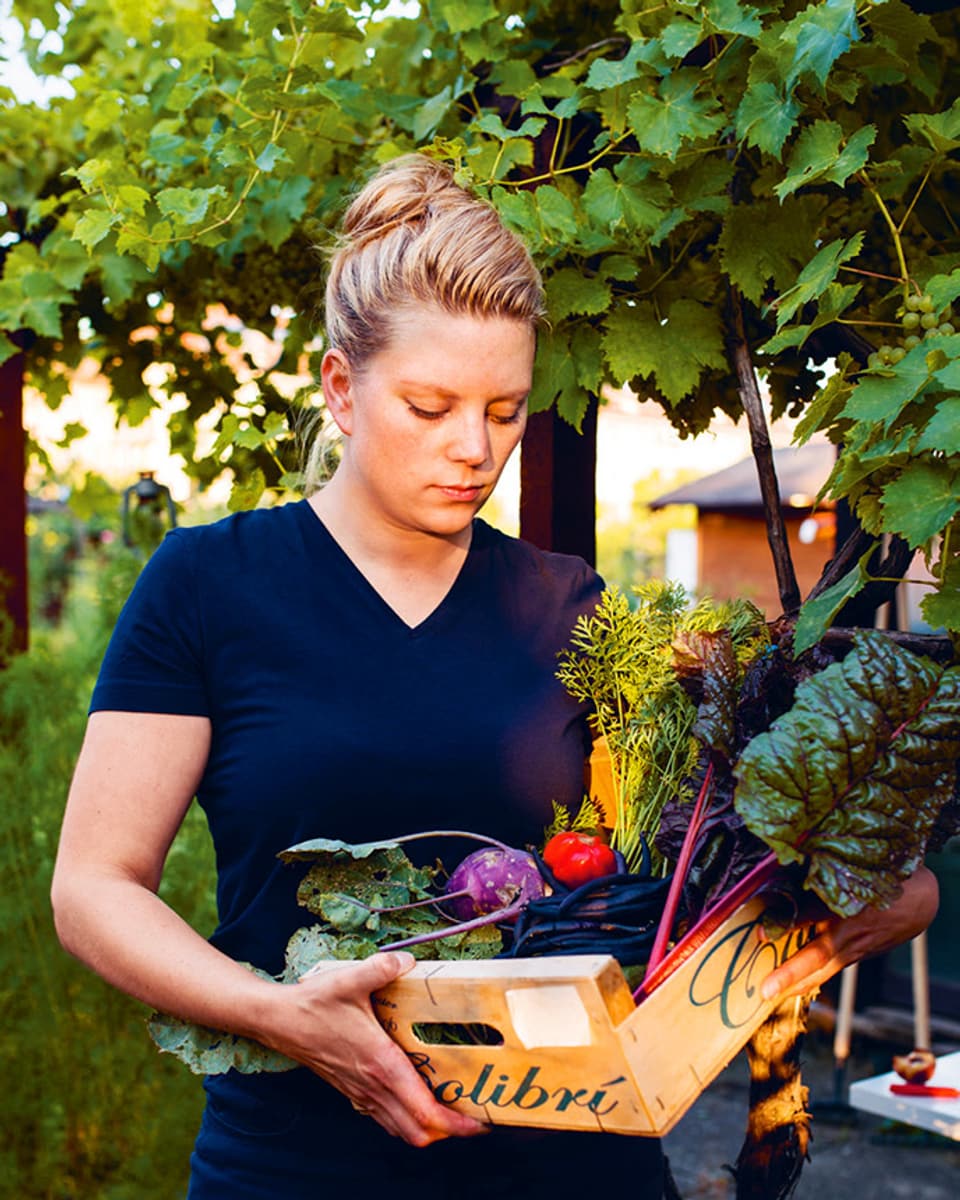 Eine junge Frau steht in einem grünen Garten und hat einen Korb in der Hand voller verschiedener Gemüse.