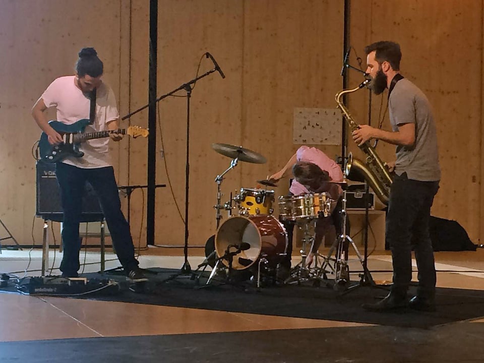 Ein Gitarrist, ein Schlagzeuger und ein Saxophonist spielen zusammen in einem Raum.