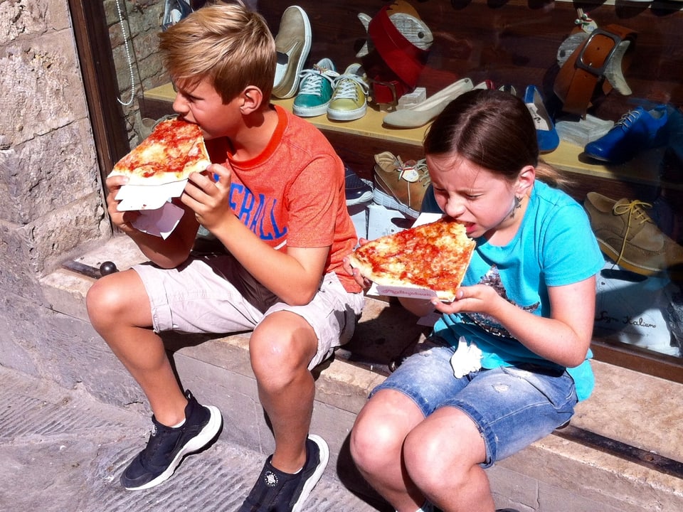Zwei Kinder essen auf Randstein je ein Stück Pizza