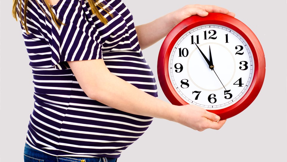Eine hochschwangere Frau hält eine Uhr, die fünf vor zwölf anzeigt. 
