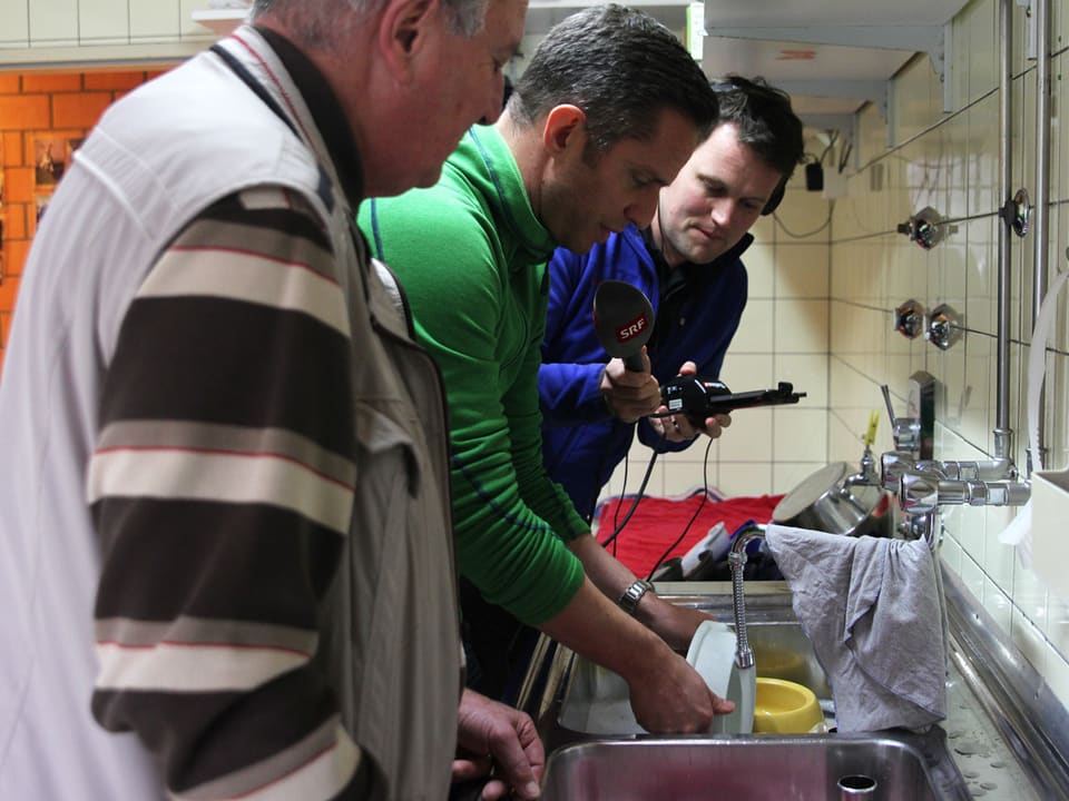 Adrian Küpfer beim Abwaschen, Ernest Schweizer und Reporter Samuel Schmid stehen neben ihm.