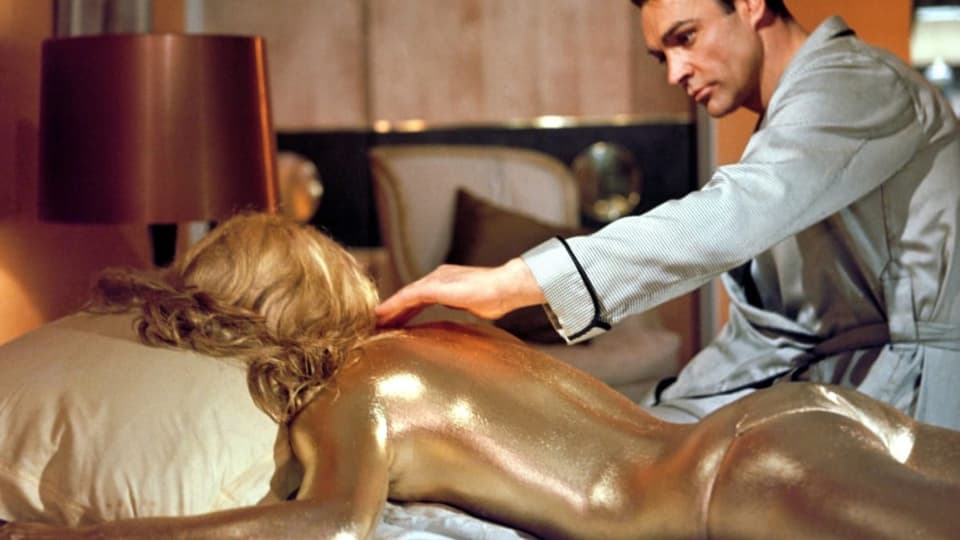 Ein Mann fasst eine im Bett liegende Frau an, deren nackter Körper vollständig mit Gold überzogen ist.