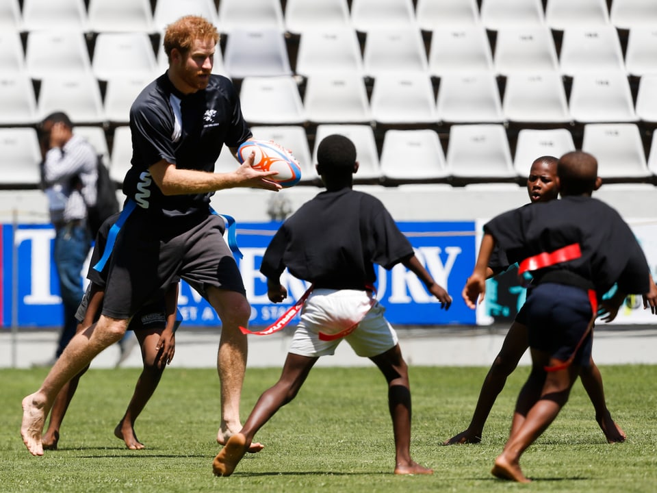 Prinz Harry hat einen Rugbyball in der Hand und spielt mit Kindern auf dem Rasen.